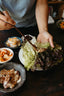 A person stands in front of a dark wood table with small plates of Korean side dishes, kimchi, and pork belly. With chopsticks, she is placing strips of cooked pork belly into curly lettuce leaves for wraps.