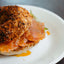 A crispy bagel sandwich with smoked salmon and fresh spring greens peeking out between the halves of the bagel. The top of the bagel is rolled in red and black flakes of spicy Red Kettle Magic Kimchi Dust. The sandwich is on a white plate with orange chili oil around the edges.