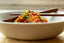 A white bowl set on a wooden table with a heaping serving of kimchi in the center, garnished with bias-cut scallions. A wooden spoon and set of chopsticks are set on top of the bowl for serving.