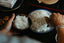 A dark, moody image of Asian-style kitchen ware on a black table. Two hands are scooping white rice with a wooden rice paddle out of a blue and white porcelain bowl into a smaller brown ceramic bowl.
