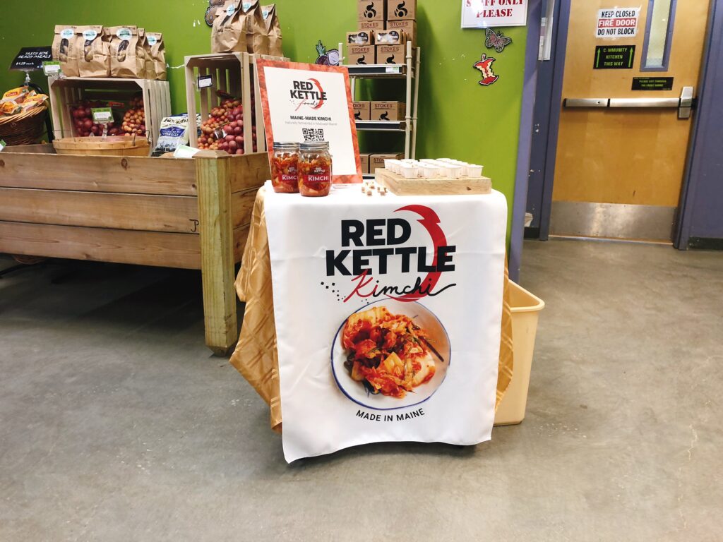 The tasting table at Rising Tide Coop in Damariscotta is set up in the produce department. It is a small table with a tablecloth reading "Red Kettle Kimchi" and product samples and jars of kimchi on top.