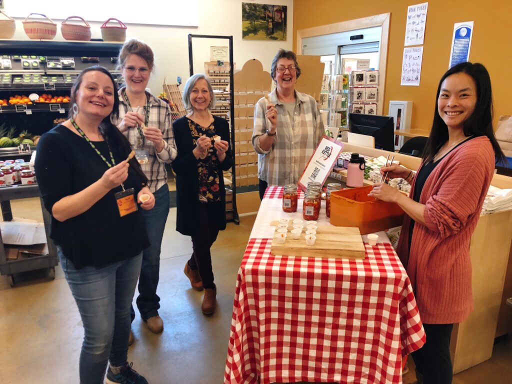 A group of employees at Blue Hill Coop sample kimchi from small cups. Ger the owner is standing behind a table covered in a checked red and white tablecloth and smiling.