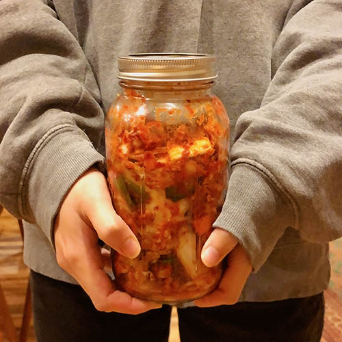 A person wearing a grey sweatshirt holds a glass quart jar full of freshly made kimchi