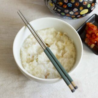 A pair of sage green and steel chopsticks rests on a white porcelain bowl with rice inside. Next to it are a lacquered teakettle and a small bowl of daikon kimchi.