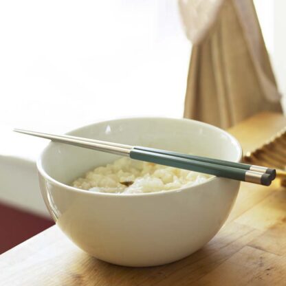 A pair of sage green and steel chopsticks rests on a white porcelain bowl with rice inside. It sits on a wooden table framed by a large window.