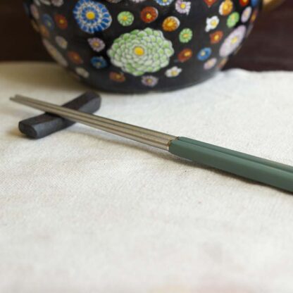 An iron chopstick rest and pair of steel chopsticks on a linen tablecloth with a lacquered teakettle in the background