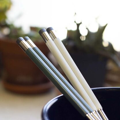 Sage green and white colored Korean chopsticks in a blue bowl, silhouetted by planters in the background