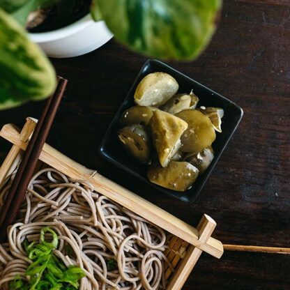 A soba noodle dish in a bamboo basket on the table next to a small black square bowl of pickles